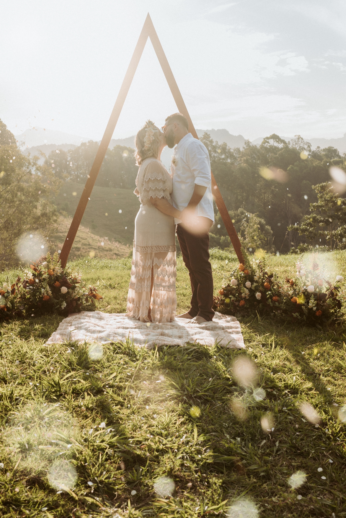 Bride wearing a Jaase dress and her groom standing in front of a triangle arbour sharing a kiss while biodegradable confetti falls around them at the hill top ceremony spot at Bunyarra in Crystal Creek NSW